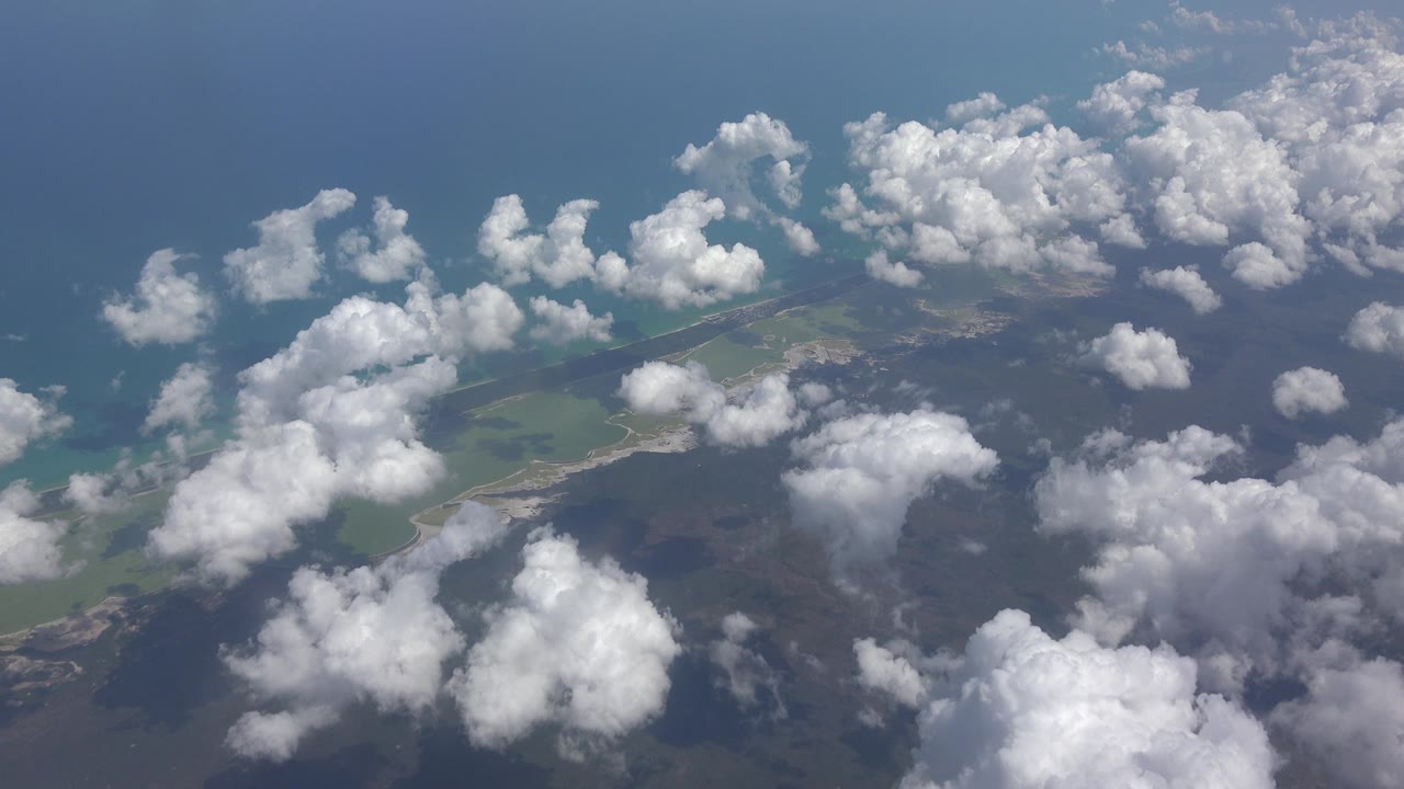 Playa Del Carmen coastline appears beneath a layered field of white clouds, with turquoise water near the shore and darker inland terrain, with no visible people.