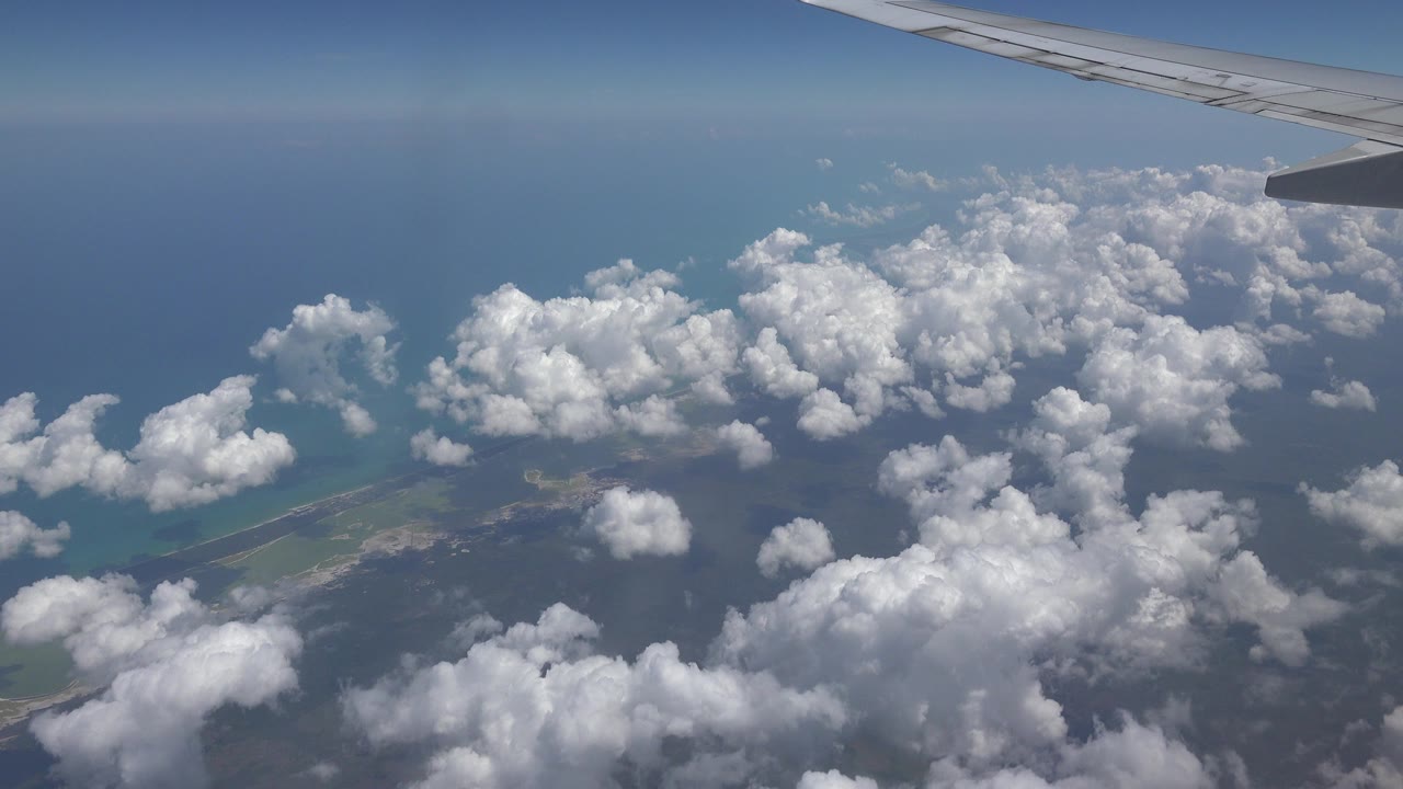 At 0:00, an airplane wing crosses the top of the frame above white clouds, blue ocean, and a thin coastline near Playa del Carmen, Mexico, with no visible people.