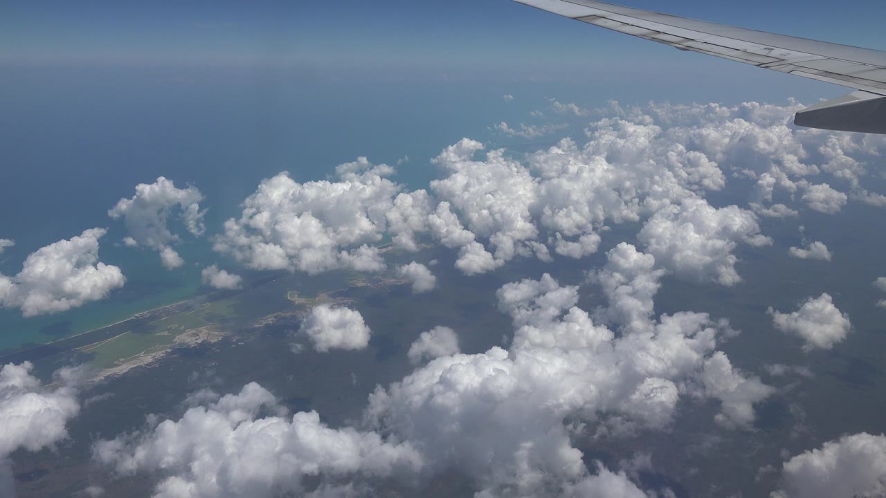 Airplane wing at the top of the frame over scattered clouds and the Playa del Carmen coastline, with blue sea beyond and no visible people.