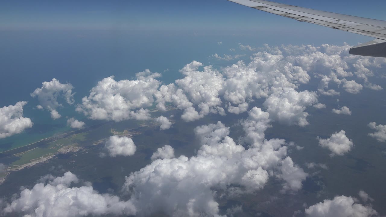 Passenger-window view over Playa del Carmen, Mexico, with an airplane wing at the top, scattered clouds over blue water, a strip of coastline at lower left, and no visible people.