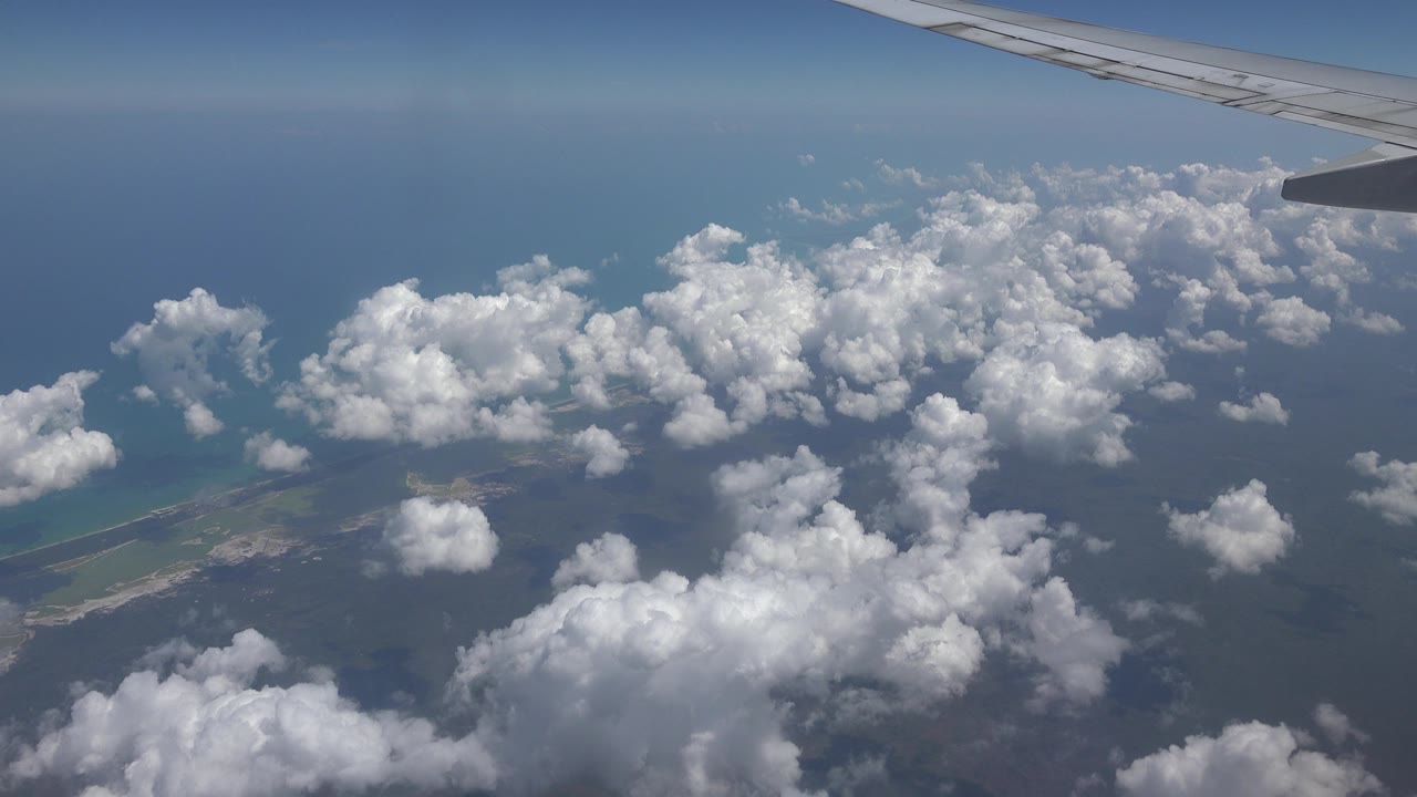 At 0:01, the airplane wing remains along the upper edge while puffy clouds cast soft shadows over water and a low coastline near Playa del Carmen, Mexico, with no visible people.