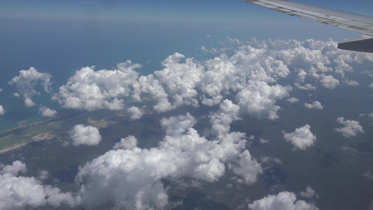 Opening frame from Playa Del Carmen, Mexico showing an airplane wing at the upper right, scattered white clouds over blue water, small coastal land below, and no visible people.