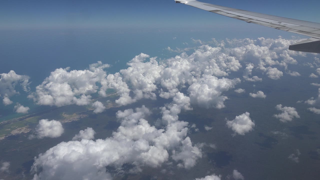 Opening view from an airplane over Playa del Carmen, Mexico, with a wing at the top right, scattered white clouds below, blue water beyond, and no visible people.