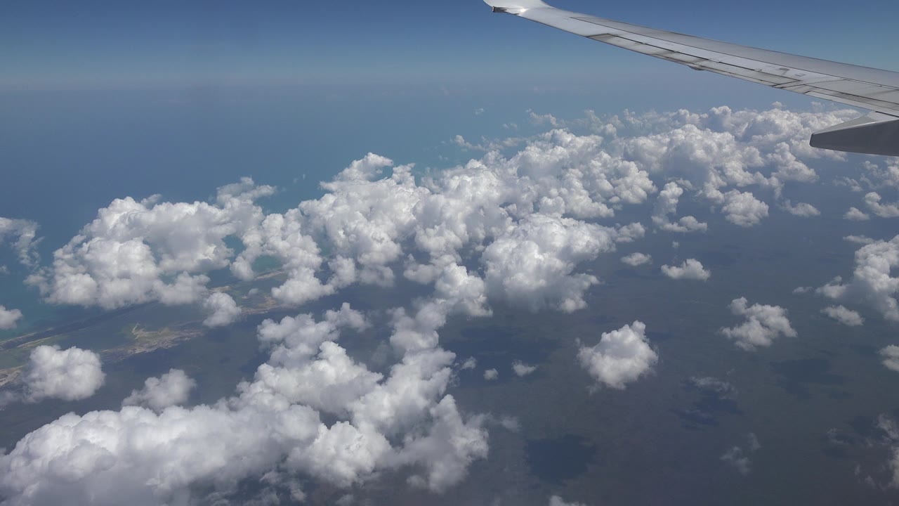 Passenger-window view over Playa del Carmen, Mexico, with an airplane wing at the top right, white clouds over blue water, and no visible people.
