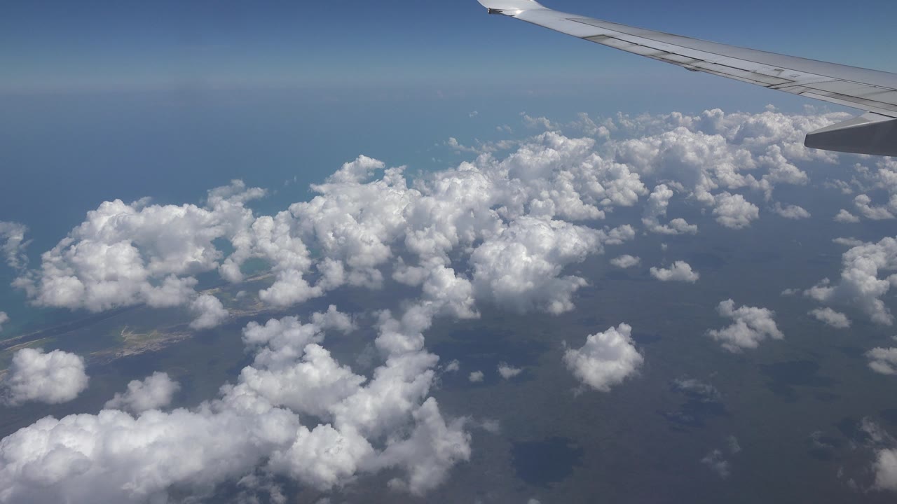 Passenger-window view over Playa del Carmen, Mexico, with the airplane wing trimmed along the upper right, a broad field of white clouds over water, and no visible people.