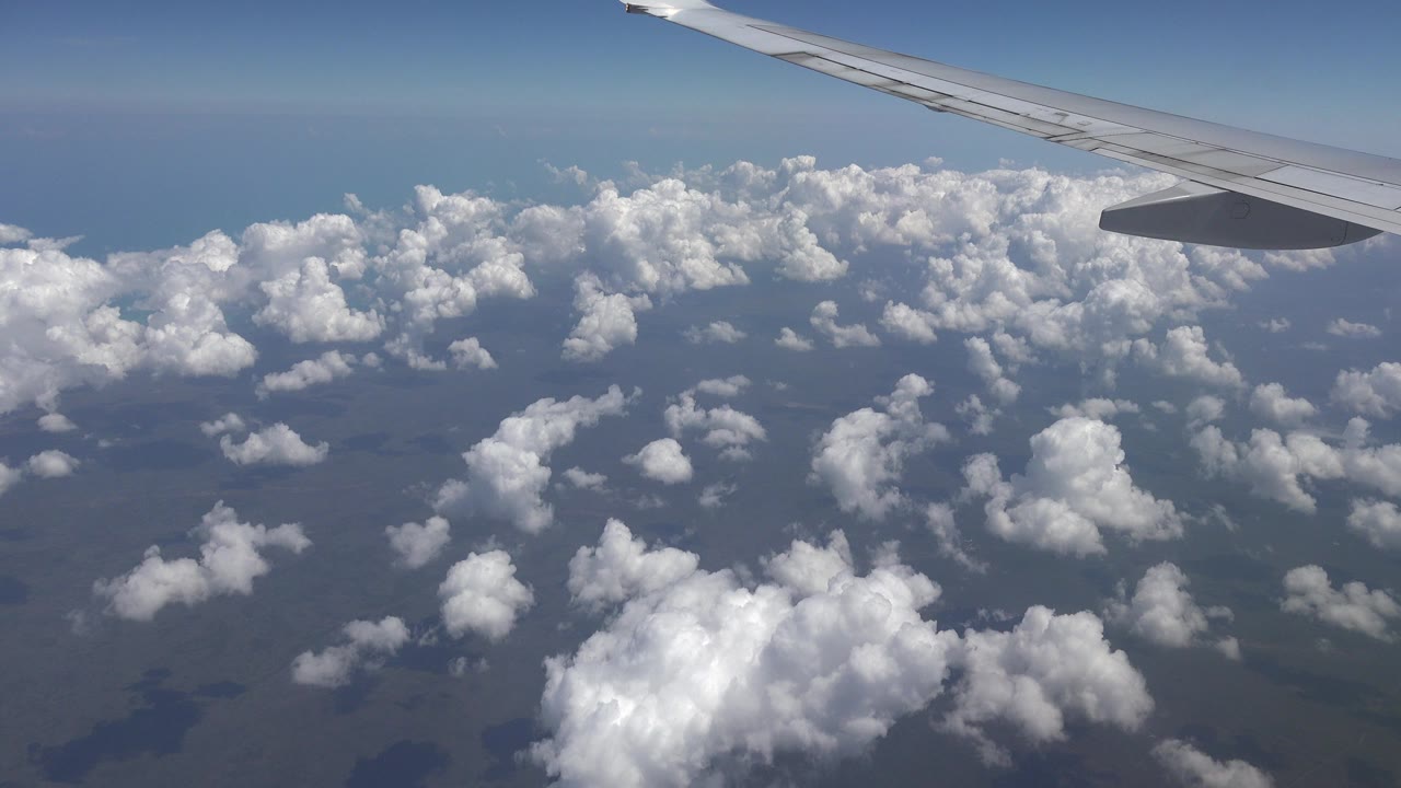 Opening frame of a Playa del Carmen, Mexico flight view with an airplane wing at the top, scattered white clouds below, blue horizon, and no visible people.