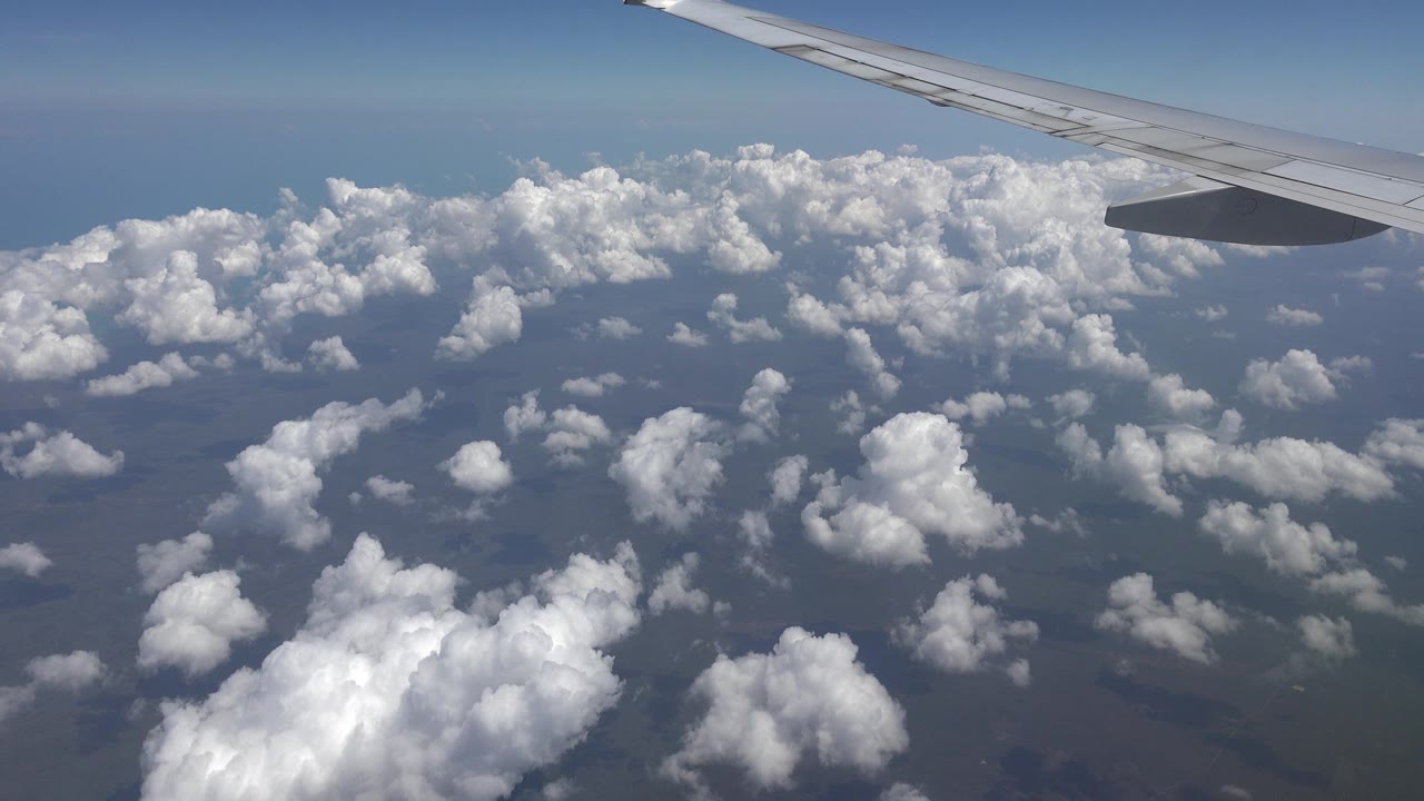 Opening view from a flying airplane near Playa del Carmen, Mexico, with the wing crossing the top of frame, bright cloud tops below, and no visible people.
