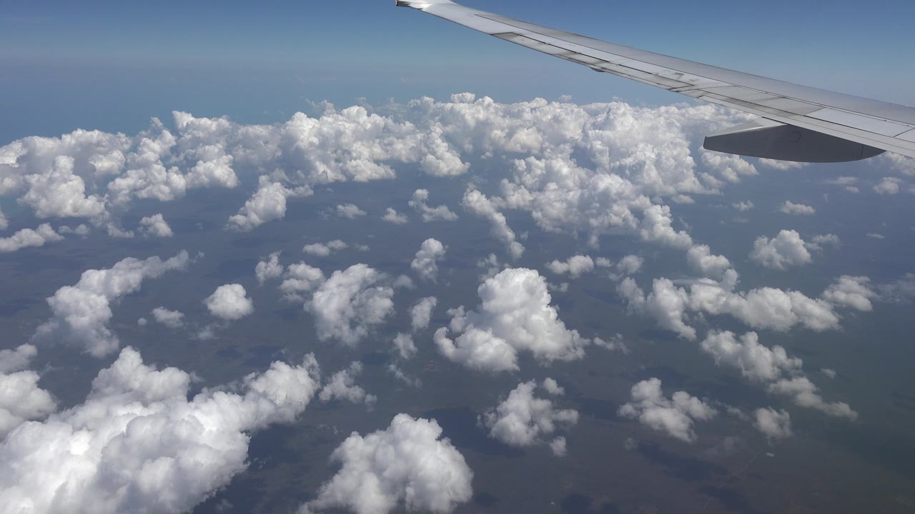 At 0:00, an airplane wing crosses the upper frame above scattered cloud tops over Playa del Carmen, Mexico, with no visible people.
