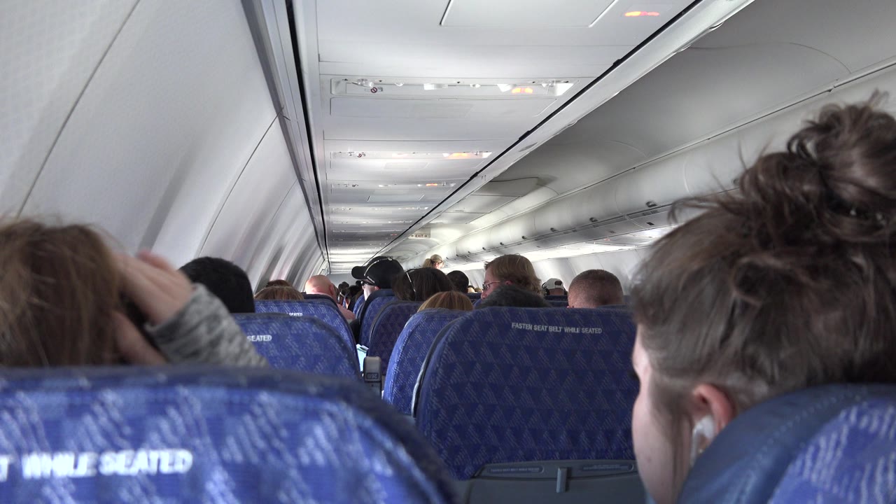 Airplane cabin seen over blue seatbacks with seated passengers ahead.
