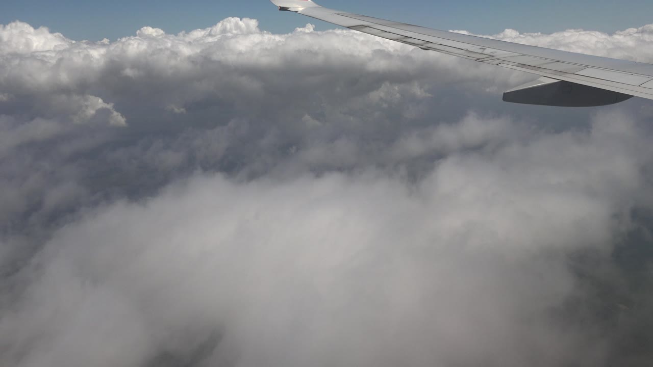 At 0:00, an airplane wing stretches across the top of the frame over thick white clouds above Playa Del Carmen, Mexico, with no visible people.