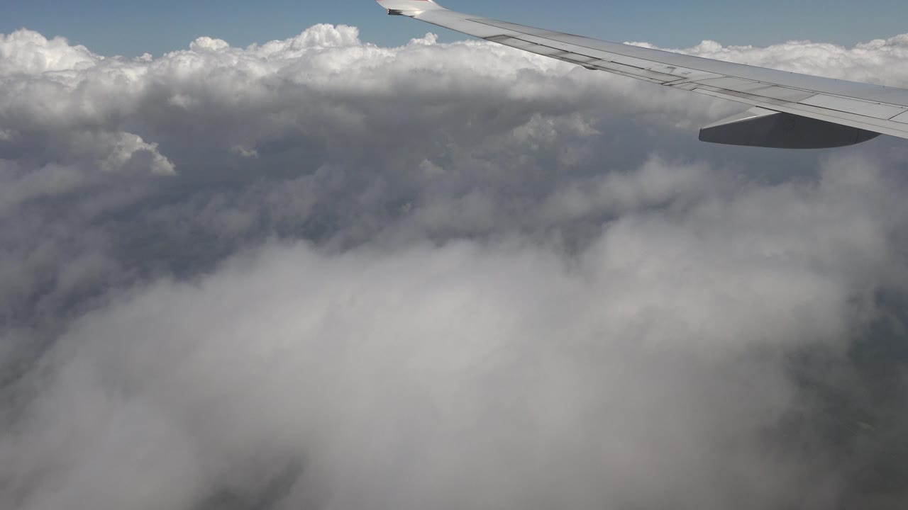 At 0:01, the airplane wing remains high in frame while layered gray and white clouds fill the view over Playa Del Carmen, Mexico, with no visible people.