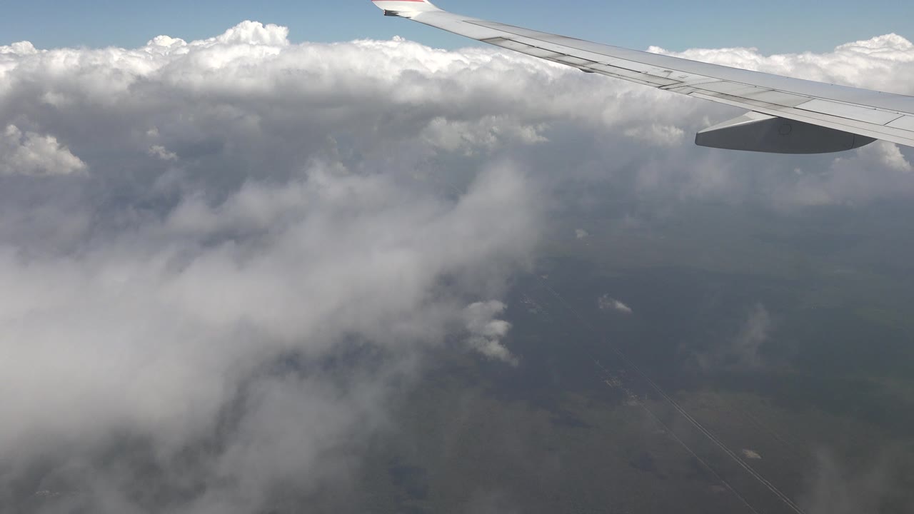 Plane wing remains near the top edge while dense white clouds fill the midground and muted terrain sits below, with no visible people.