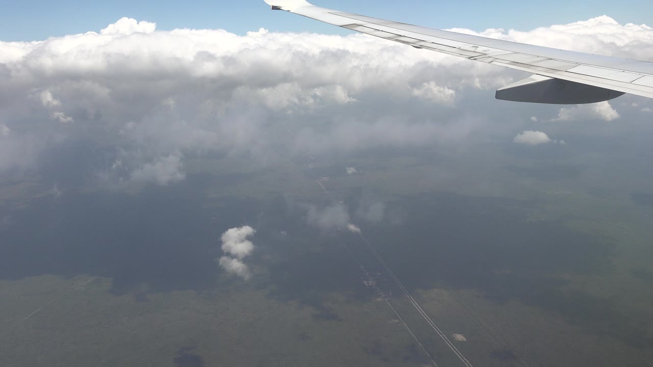 Airplane wing seen from a passenger window over Playa del Carmen, Mexico, with bright clouds, blue sky, hazy ground below, and no visible people.