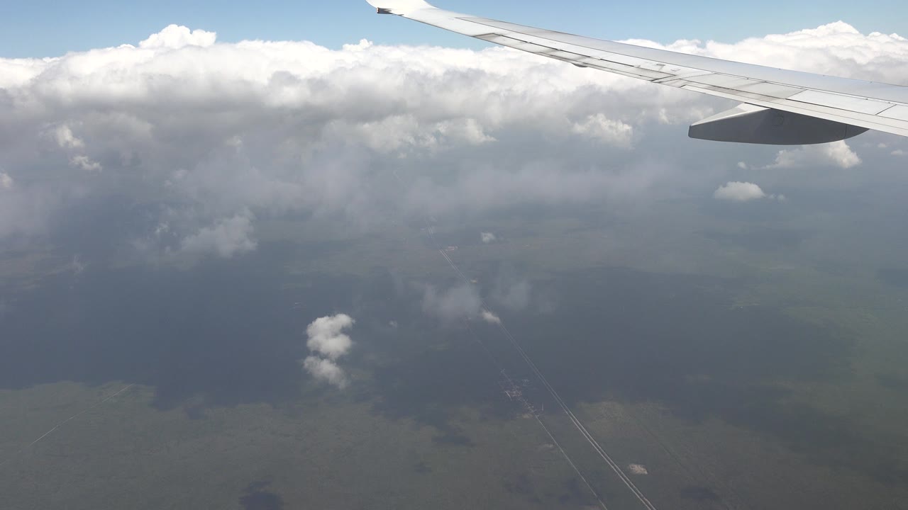 Passenger-window view over Playa del Carmen, Mexico, with the aircraft wing near the top, a wide cloud bank, muted ground, and no visible people.