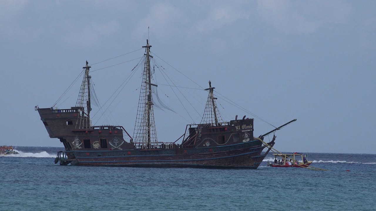 Wide view of a large dark sailing-style ship offshore in Playa del Carmen, Mexico, with a small boat at the right edge, breaking surf behind, and no visible people.