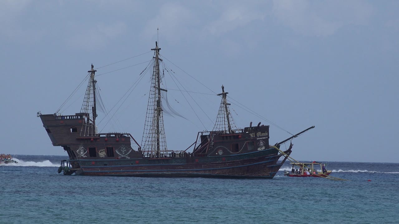 The ship remains centered off Playa del Carmen, Mexico, with tall masts, rigging, blue foreground water, a small boat to the right, and no visible people.