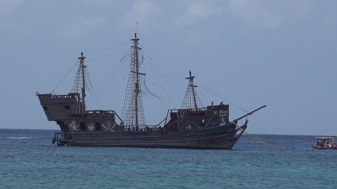 Dark sailing ship on blue-green sea with a small tugboat nearby.