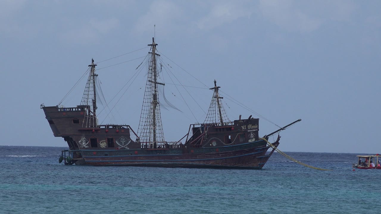 At 0:01 in Playa del Carmen, Mexico, the sailing ship remains broadside on the sea as the small tugboat sits near the right edge and the tow line angles forward, with no visible people.