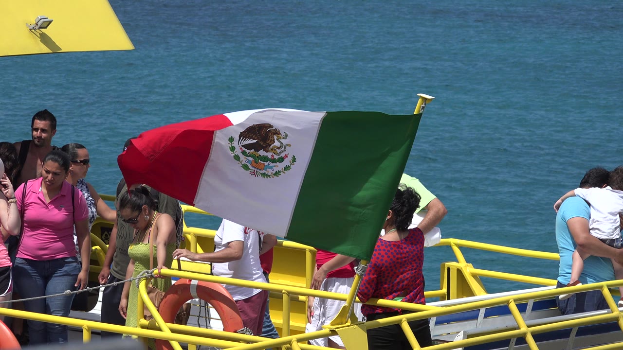 A Mexican flag in Playa del Carmen waves across a yellow boat deck while passengers stand nearby and blue water fills the background.