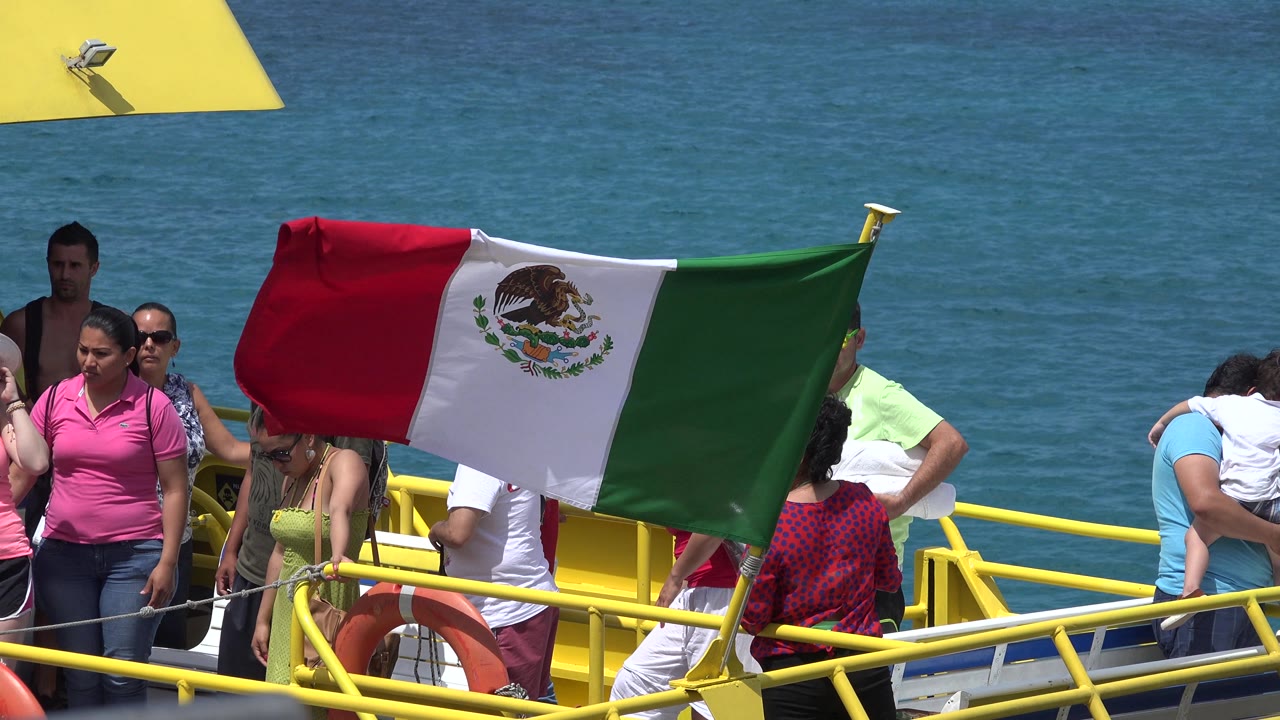 Mexican flag raised above passengers on a yellow boat against blue water.