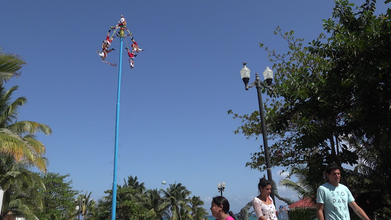 In Playa del Carmen, Mexico, the performers remain high against the blue sky while a man and woman pass below near a leafy tree and twin street lamps.