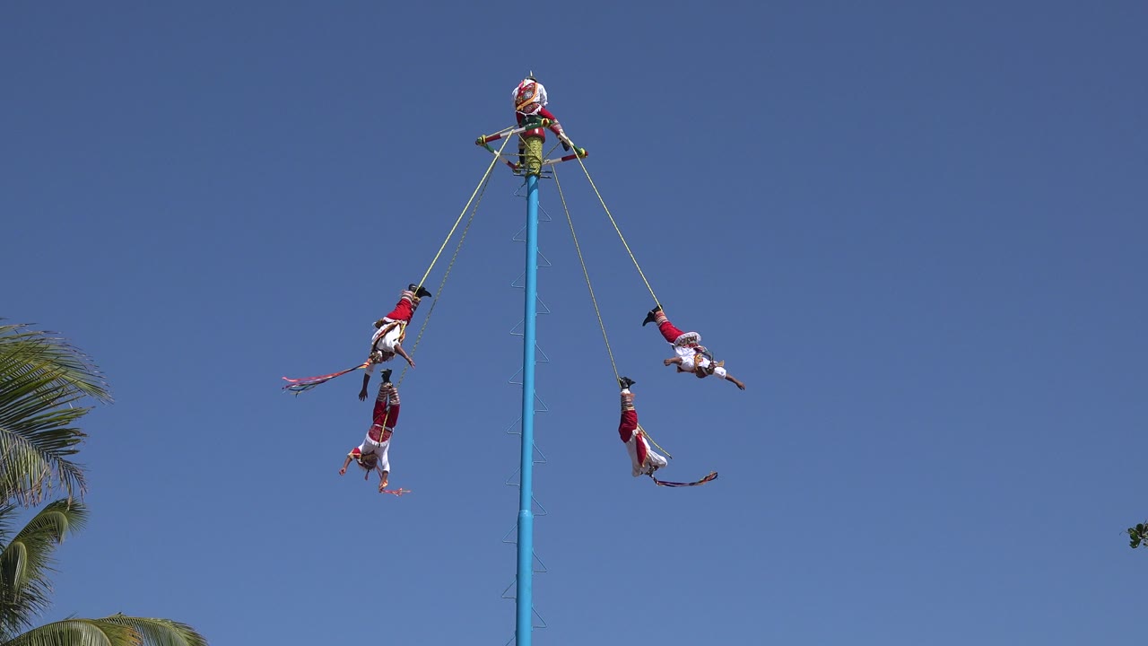 In Playa del Carmen, Mexico, several suspended people are spaced around a tall blue pole as the ropes angle outward from the platform near the top.
