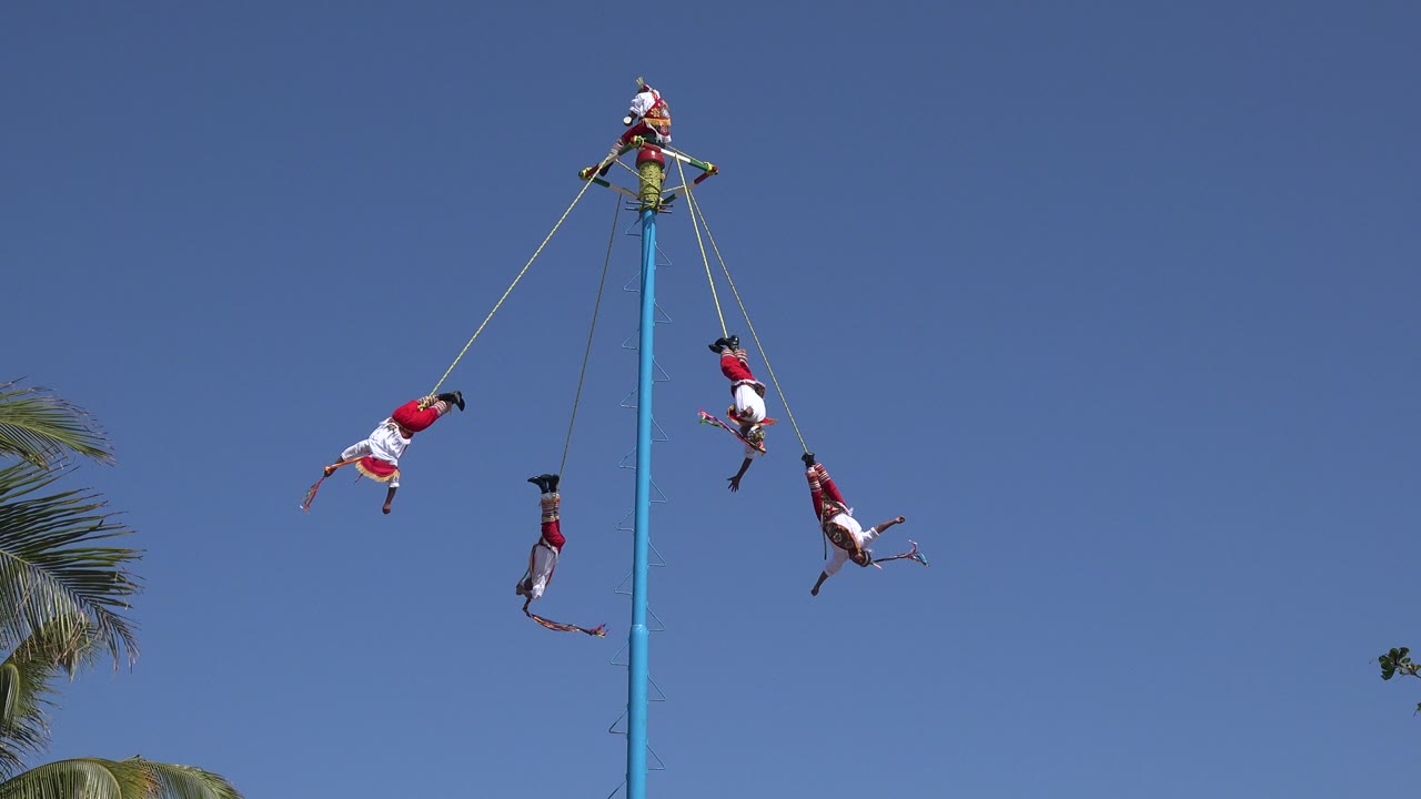 People on ropes rotate near the top of a blue pole.
