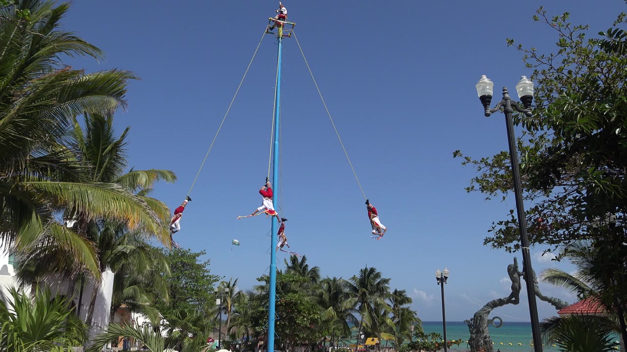 In Playa del Carmen, Mexico, multiple performers in red and white hang from ropes around a tall blue pole, with palm trees, street lamps, blue sky, and distant water behind them.