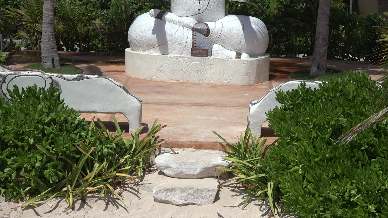 At 0:01, the camera view holds on the statue's crossed legs and round base in Playa Del Carmen, framed by bright green shrubs and no visible people.