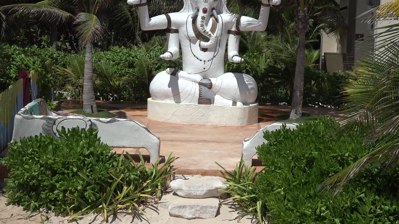 White elephant-headed sculpture on a platform behind shrubs and palm trees.