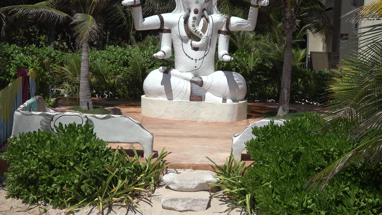 The Playa del Carmen sculpture remains partly cropped as palms and dense greenery fill the background, with the low white wall and a partially visible person at the left edge.
