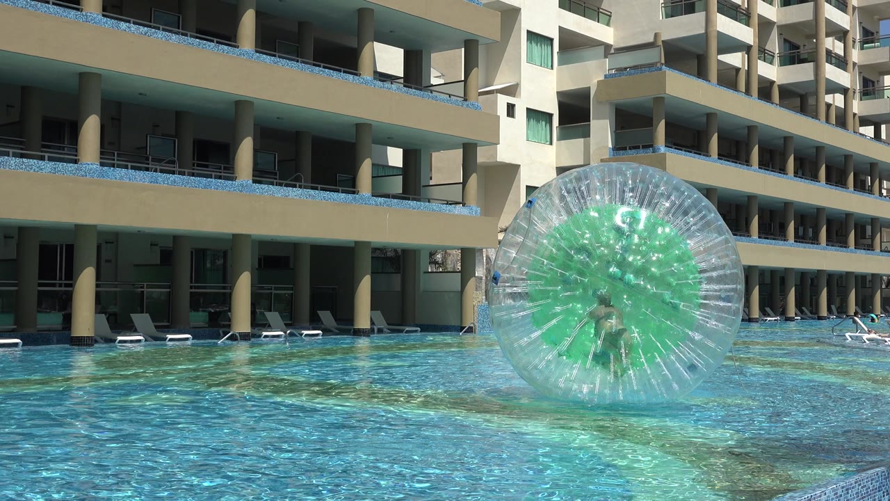 Person inside a clear inflatable ball on a blue pool by modern buildings.
