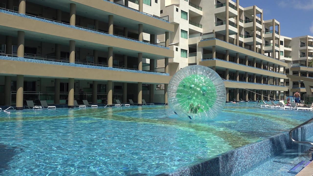 At 0:01 in Playa del Carmen, Mexico, the inflatable ball sits farther across the pool surface while the apartment building and pool ladder remain visible.