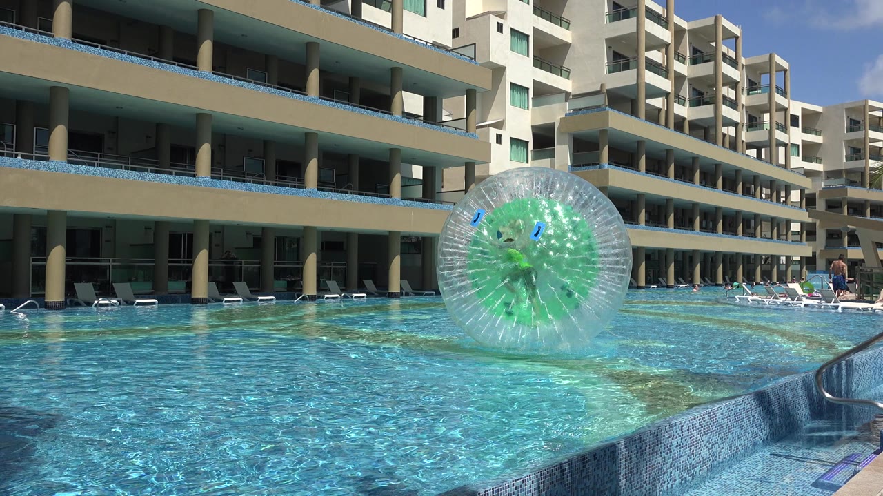 A transparent green-centered water-walking ball with a person inside sits left of center on a bright pool in Playa Del Carmen, Mexico, with modern buildings behind it.