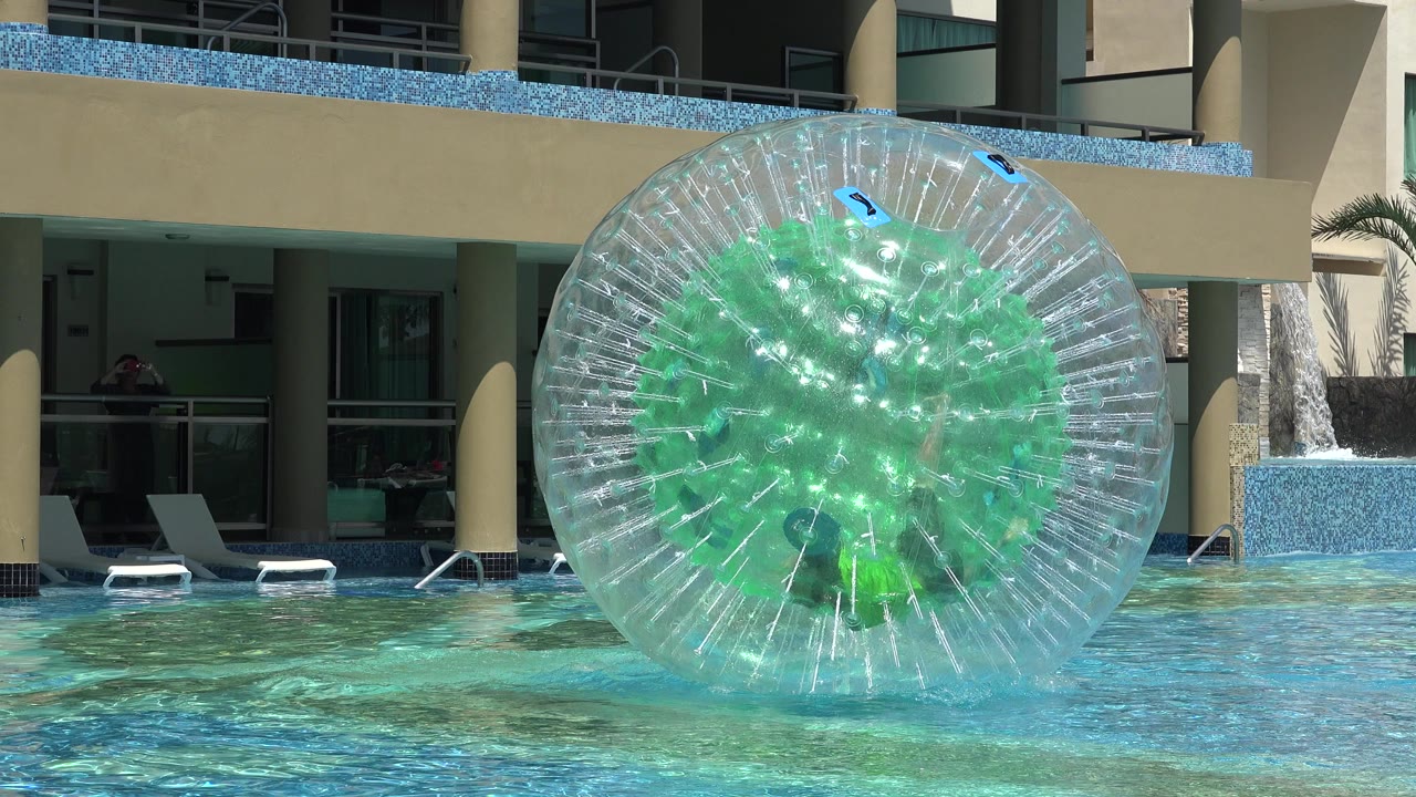 A boy is partly visible inside a clear green-centered inflatable ball floating in a Playa Del Carmen swimming pool, with lounge chairs and resort balconies behind it.