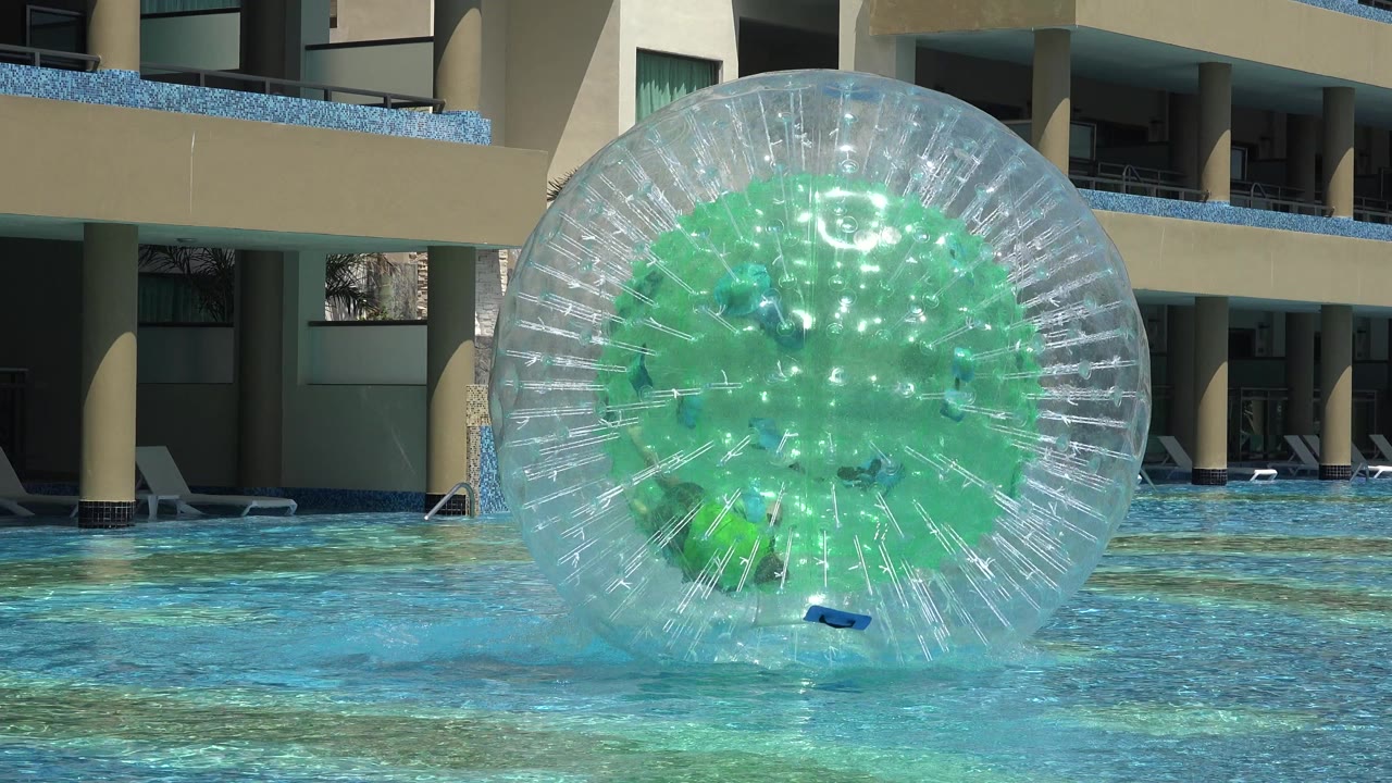 A clear inflatable water ball with green panels floats near center frame in a Playa del Carmen hotel pool, with a person partly visible inside and balconies behind it.