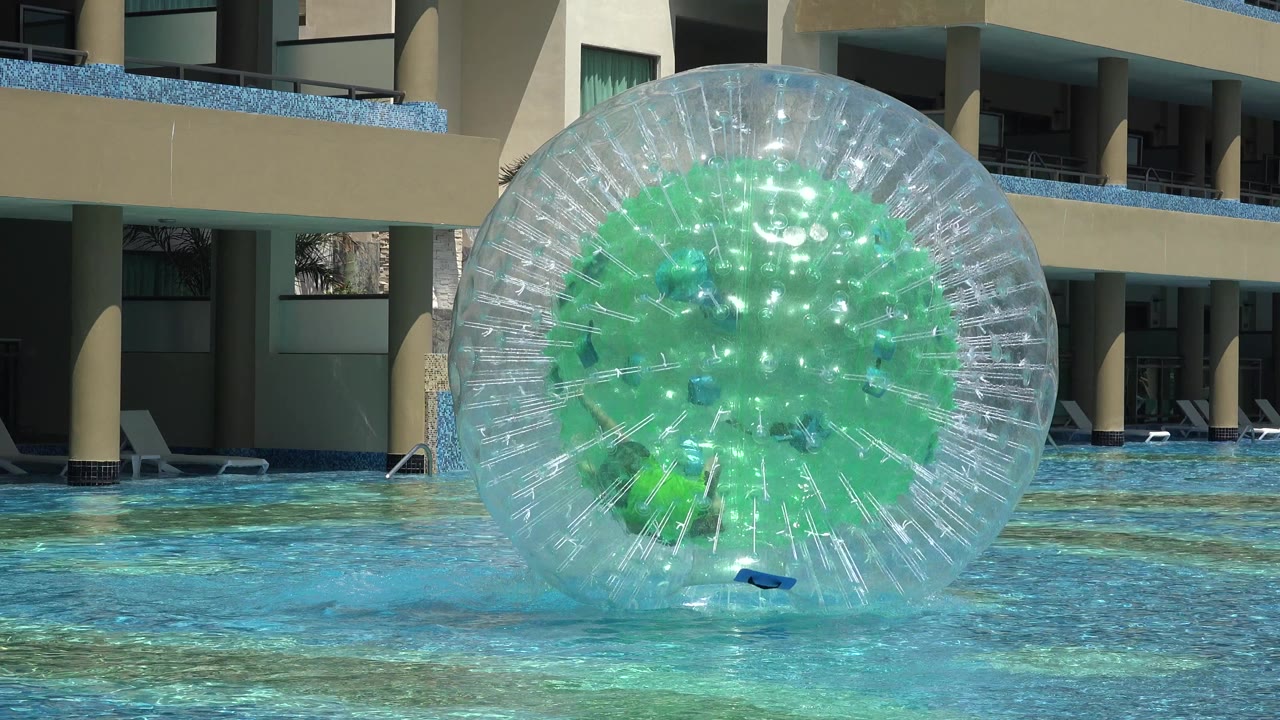 The inflatable water ball remains centered in the Playa del Carmen pool, with the person inside obscured by reflections and green panels while sunlight glints on the plastic surface.