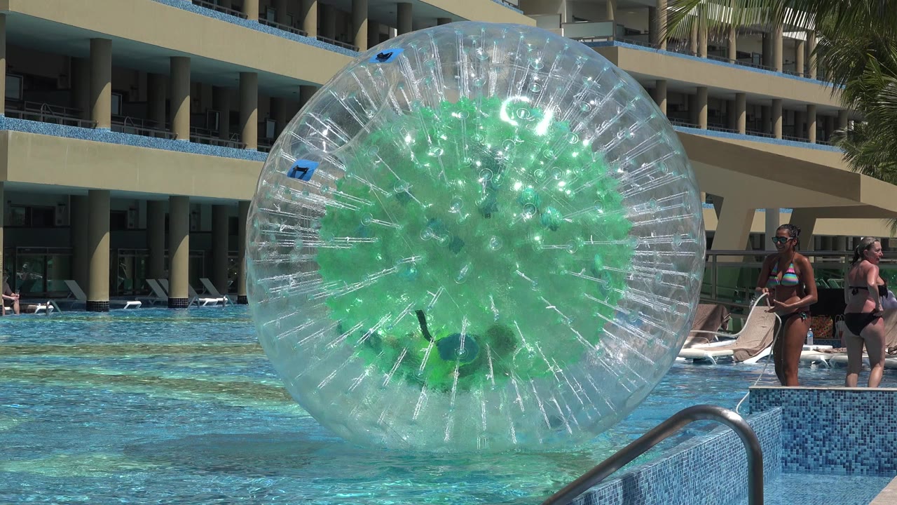 The inflatable water ball sits centered in the Playa del Carmen hotel pool, with two adult guests on the deck and stacked balconies in the background.