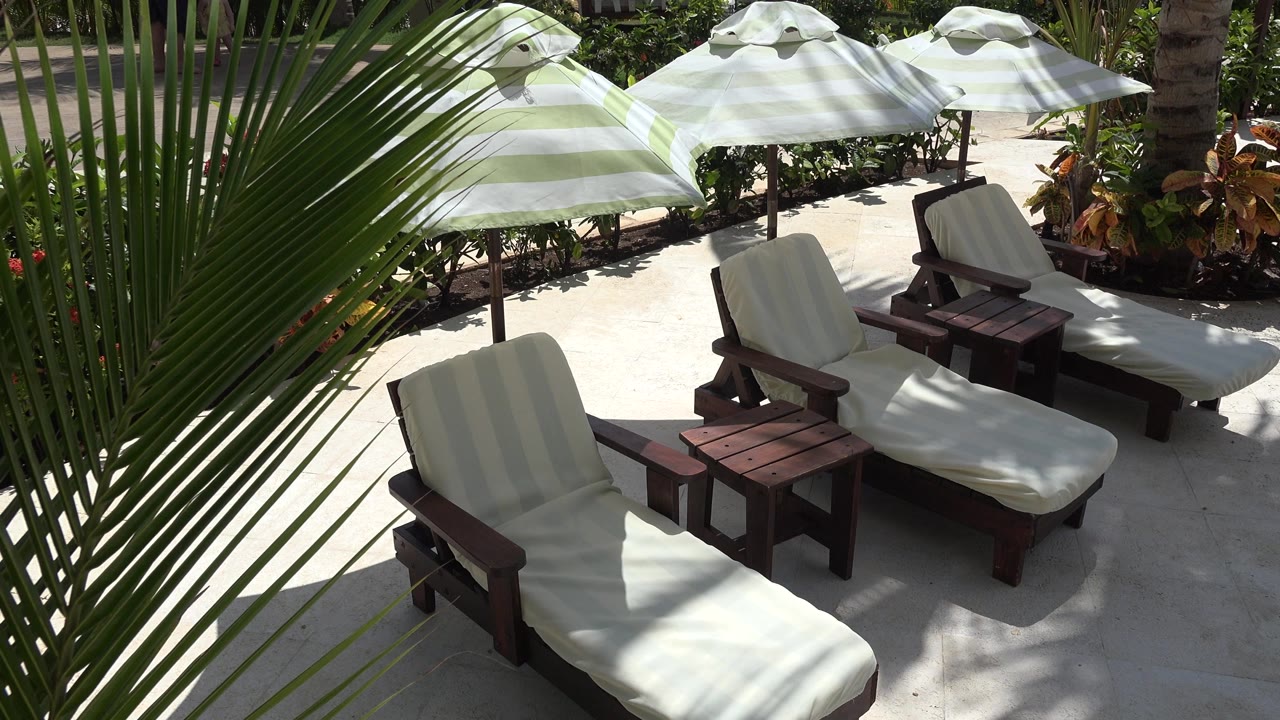 Empty lounge chairs and dark wooden side tables sit on a bright terrace in Playa del Carmen, Mexico, with umbrellas casting striped shade and no visible people.