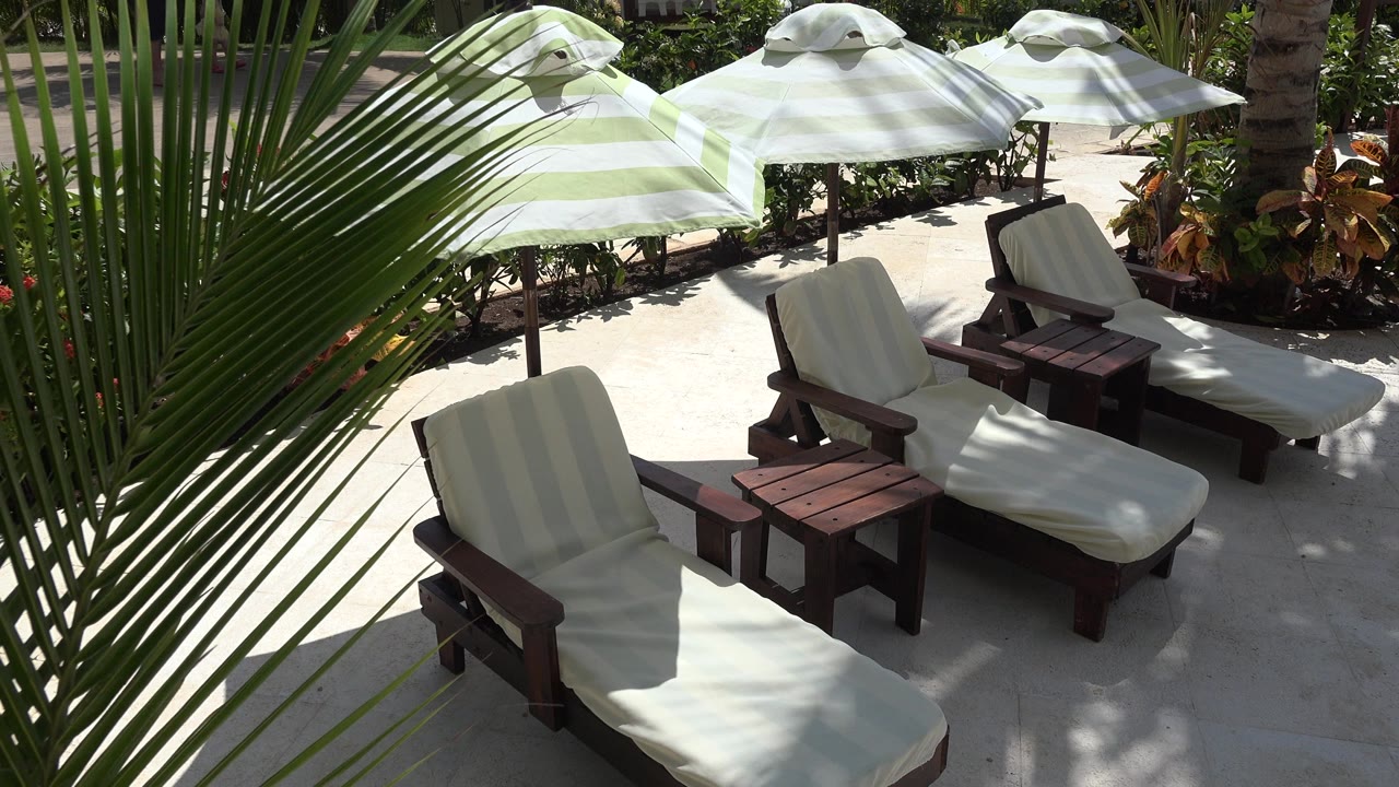 Empty lounge chairs with light cushions sit beside dark wooden tables beneath striped umbrellas in Playa Del Carmen, Mexico, with dappled sun on the patio and no visible people.