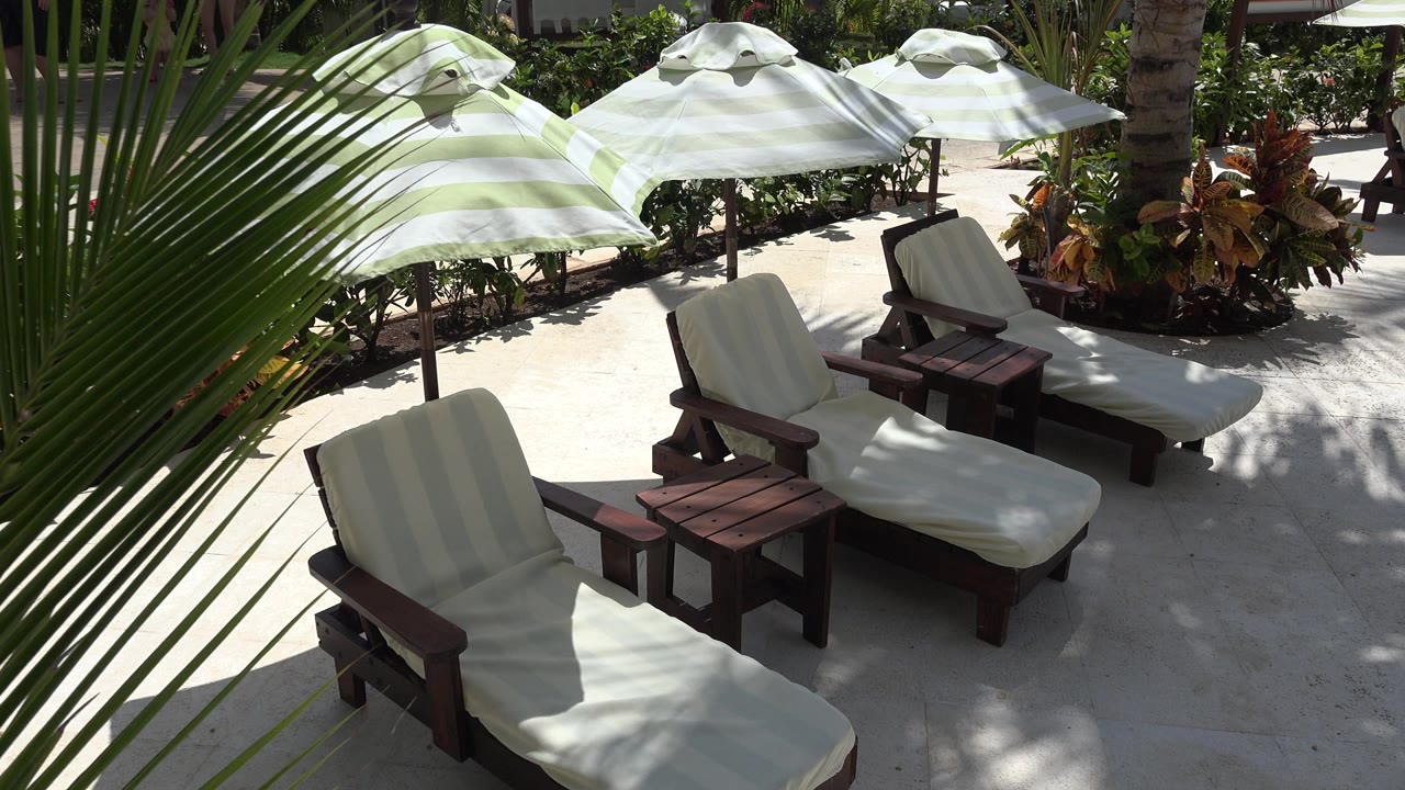 At 0:01, the same Playa del Carmen cabana area shows two cushioned lounge chairs, dark wooden tables, pale striped umbrellas, dense planting, and no visible people.