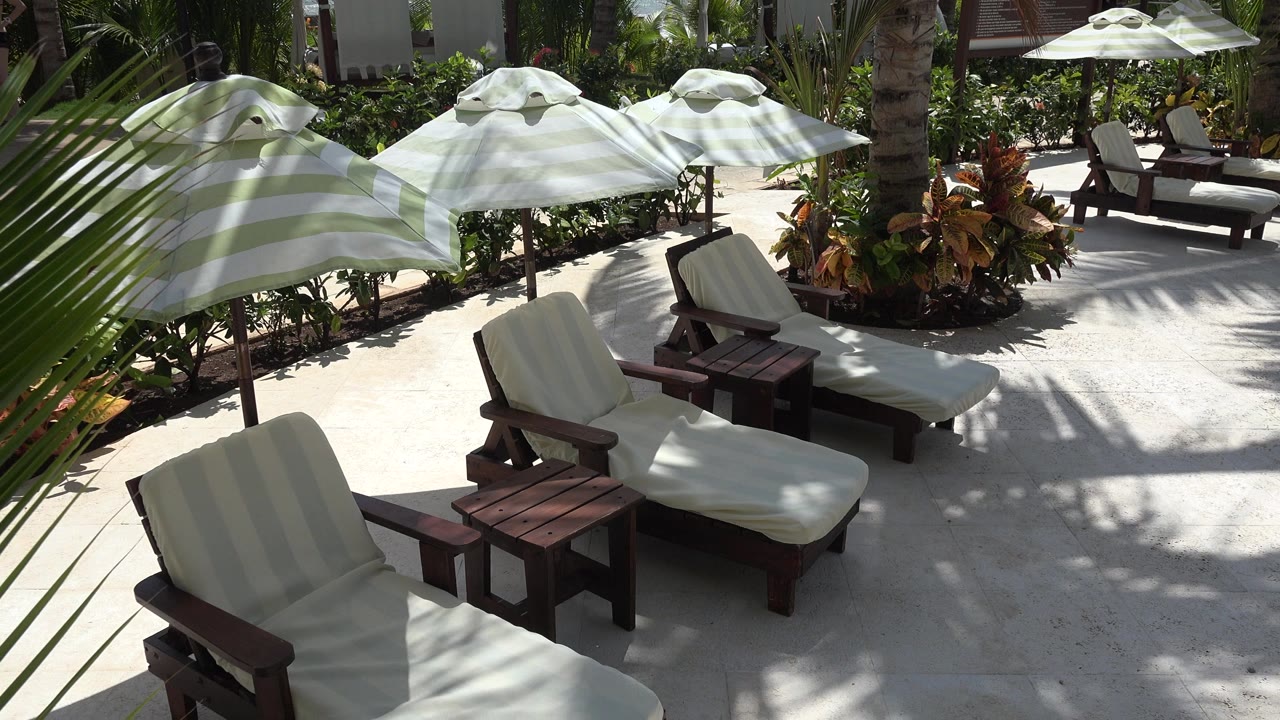 Opening frame of empty white-cushioned lounge chairs and striped umbrellas on a sunlit Playa del Carmen patio with tropical plants and no visible people.