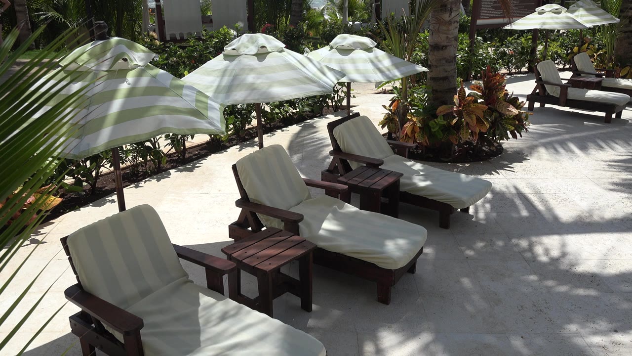 Second frame showing the same empty Playa del Carmen lounge area, with palm fronds crossing the foreground and white umbrellas shading brown chairs with no visible people.