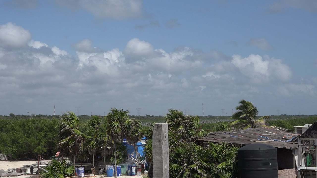 Opening frame of Playa Del Carmen, Mexico, showing palms, rough rooflines, a black water tank, sandy ground, and a cloudy blue sky with no visible people.