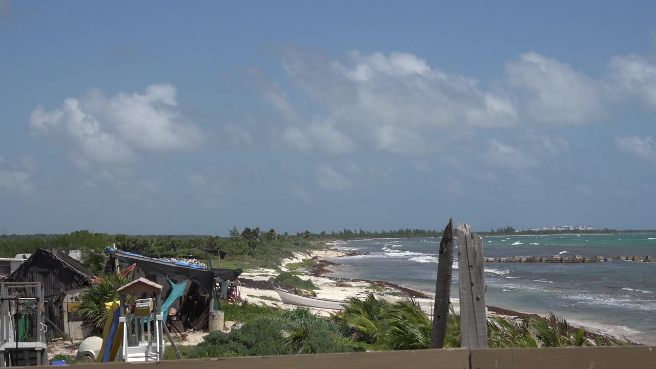 A bright Playa del Carmen beach frame with wind-bent palms, turquoise shallows, white surf, and rustic shorefront structures in the foreground.