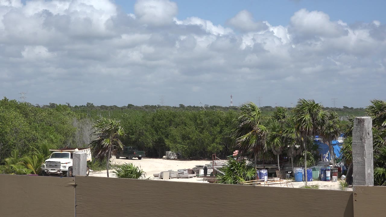 Playa del Carmen rooftop view with the white truck at left, construction materials on pale sand, palms at center, green foliage behind, and no visible people.