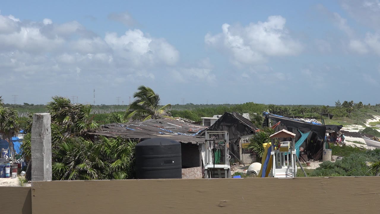 Playa del Carmen coast with rough-roofed shore buildings, a small slide-like structure, palms in wind, beach, ocean waves, and small people visible near the midground.