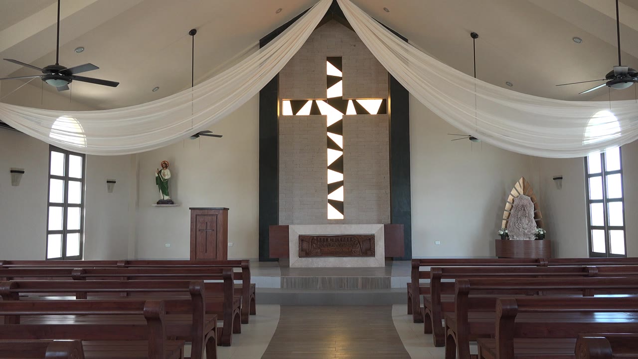 Opening frame of an empty Playa Del Carmen church-like interior with wooden pews leading toward a black and white geometric cross, ceiling drapery, tall windows, and no visible people.