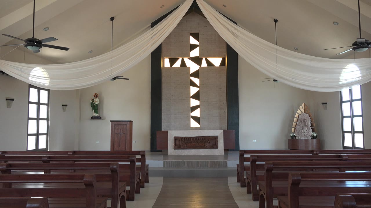 Second frame holds on the Playa Del Carmen interior as the pew rows, altar platform, tall side windows, ceiling fans, and central patterned cross remain visible with no visible people.