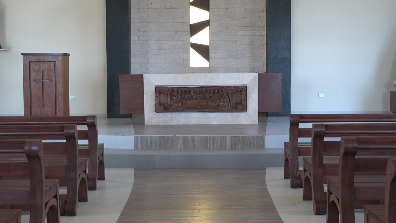 Opening frame of a Playa Del Carmen church interior with wooden pews at left and right, a raised altar area, pale walls, and no visible people.